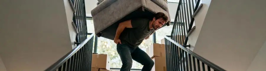 Homme transportant seul canapé dans escalier, effort intense et posture dangereuse lors déménagement solo