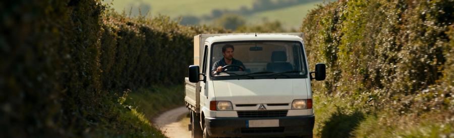 Camion déménagement manœuvrant sur chemin rural étroit bordé végétation dense, accès difficile campagne